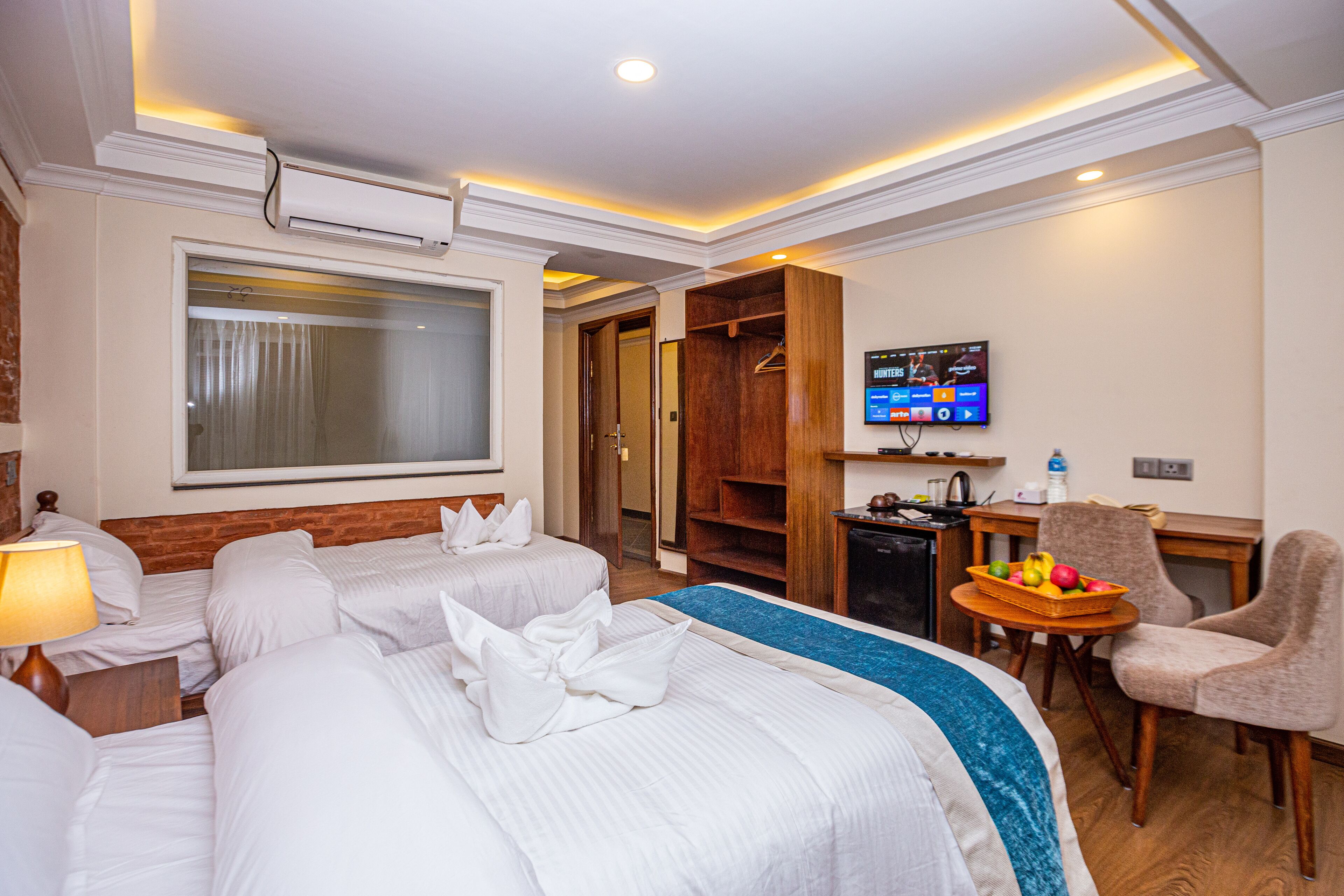 A hotel room with two beds, a TV, a desk with a fruit basket, and a chair.