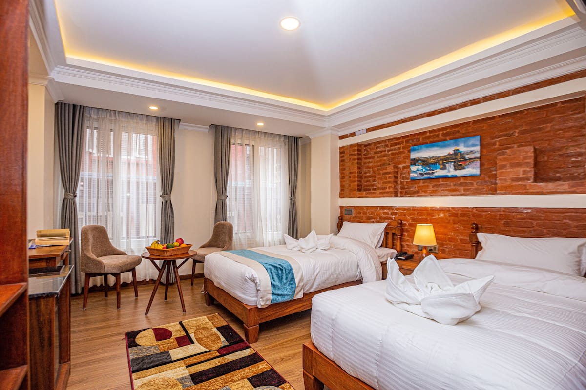 Habitación de hotel con dos camas, un escritorio, una silla, una mesita con fruta y un cuadro en la pared de ladrillo.