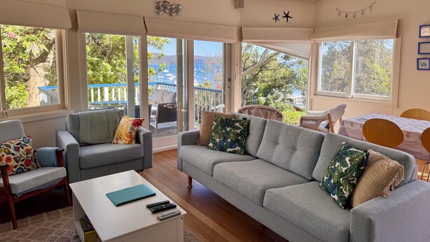 Ein geräumiges Wohnzimmer mit grauem Sofa, Sesseln und einem Couchtisch. Große Fenster bieten Blick auf das Meer und Boote.