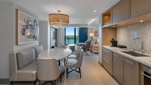 A modern hotel room with a dining area, a kitchen with marble countertops, and a large window offering a view of the sea.