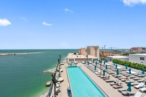 A rooftop pool with sun loungers and umbrellas, overlooking a body of water and a cityscape.