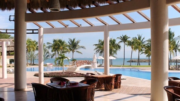 A resort pool area with lounge chairs and umbrellas, overlooking a clear blue sea and palm trees.