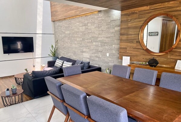 A modern living room with a large wooden dining table, grey upholstered chairs, a black sofa with cushions, a flat-screen TV mounted on the wall, and a wooden accent wall.