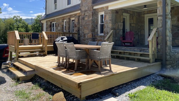 A wooden deck with outdoor furniture, a stone pillar, and a covered porch area. - River Stone Farmhouse on 35 acres with trout stream (Andrews)