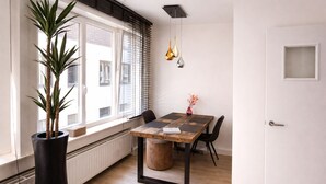A dining area with a wooden table, chairs, a potted plant, and a window with blinds. - Oostende Luxe Appartement in het Centrum Voor 2 +2 (Oostende)