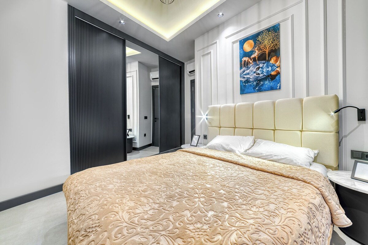 A modern bedroom with a large bed, a nightstand, a wall-mounted lamp, and a painting above the bed.