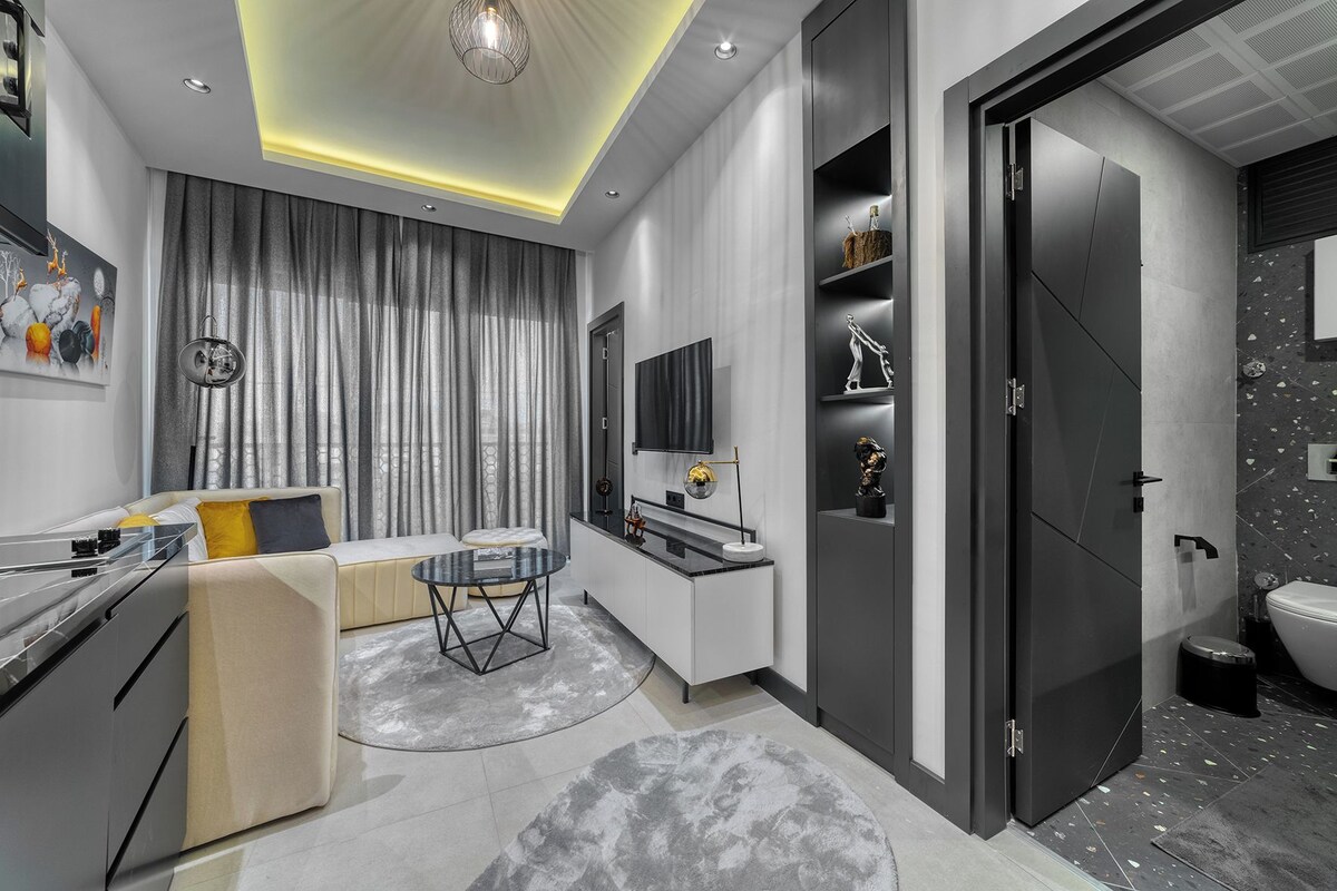 A modern living room with a sofa, a coffee table, and a TV. There is a kitchen area with a countertop and cabinets.