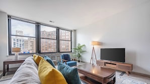 A modern living room with a sofa, armchairs, a coffee table, a TV, and a large window with a city view.