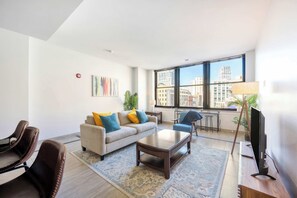 A modern living room with a sofa, coffee table, and chairs, a TV, and a large window with a city view.