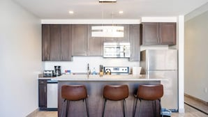A modern kitchen with dark cabinets, a stainless steel refrigerator, a microwave, and a coffee maker. There are three bar stools at the island.