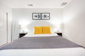 A hotel room with a bed, two bedside lamps, a gray checkered throw, and two abstract wall art pieces.