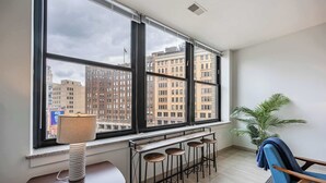A room with a large window, a desk with a lamp, a bar stool area, and a blue chair.