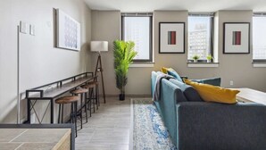 A modern living room with a grey sofa, a coffee table, and a bar area with stools. There are framed artworks on the walls and a large window providing a view of the city. - 2BR Executive Modern Downtown Apartment (Detroit)