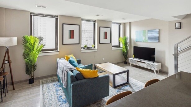 A modern living room with a sofa, coffee table, and TV. There are large windows, a staircase, and a plant. - 2BR Executive Modern Downtown Apartment (Detroit)