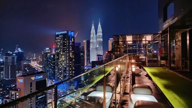 Een dakterrasbar met uitzicht op de stad, waaronder de Petronas Towers, en een glazen hekwerk dat uitkijkt over het stadslandschap.