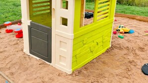 A small, colorful playhouse with a red roof and green walls, situated on a sandy area with toys scattered around. - Extertal-ferienpark - Komfort-ferienhaus Wandern Mountainbike Sauna #50 (Extertal)