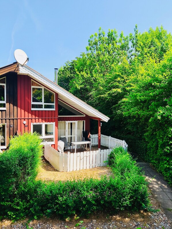 Une maison rouge avec une clôture blanche, une terrasse dotée de mobilier d’extérieur et une végétation luxuriante.