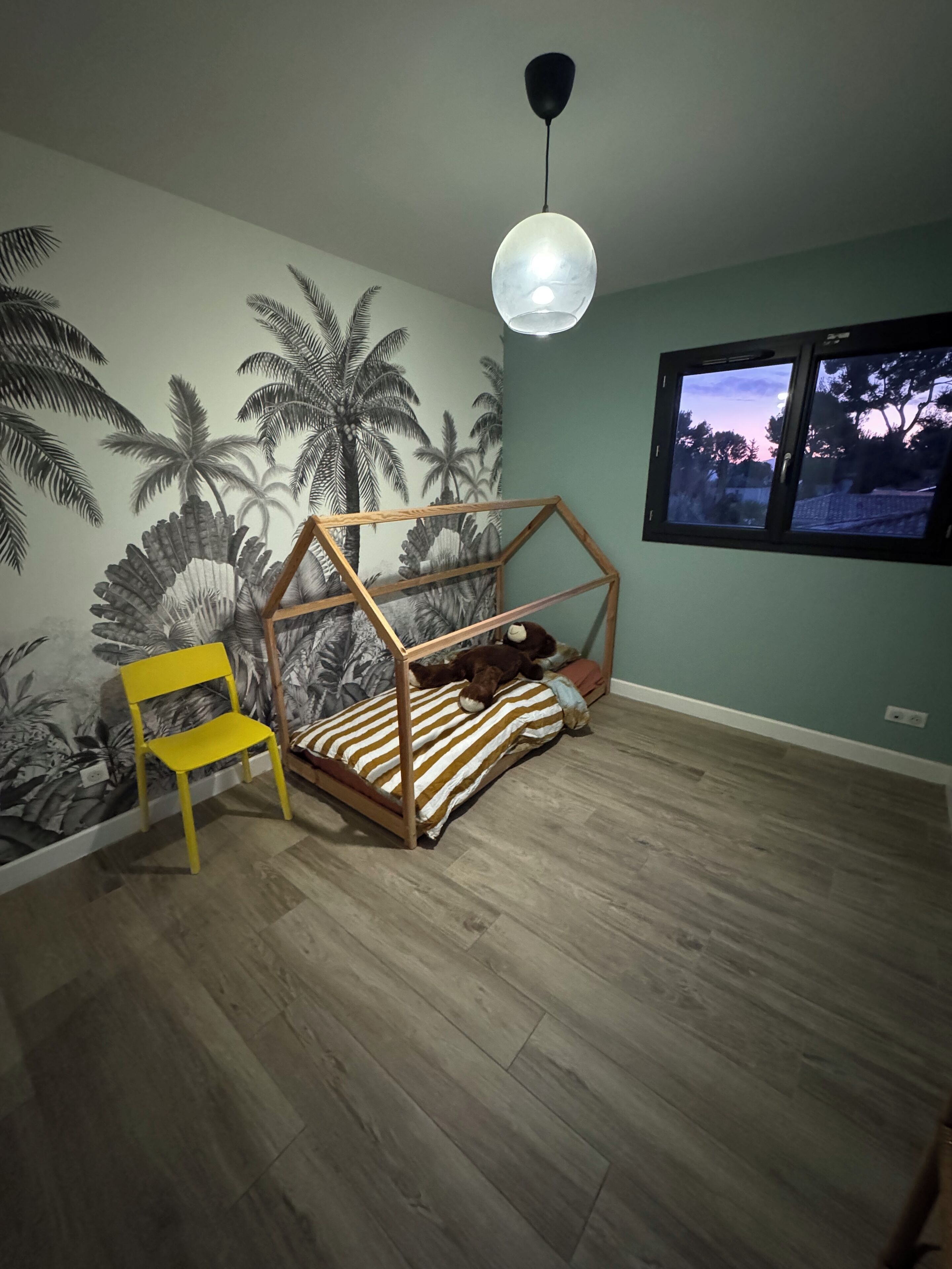 A room with a bed, a chair, and a mural of palm trees.