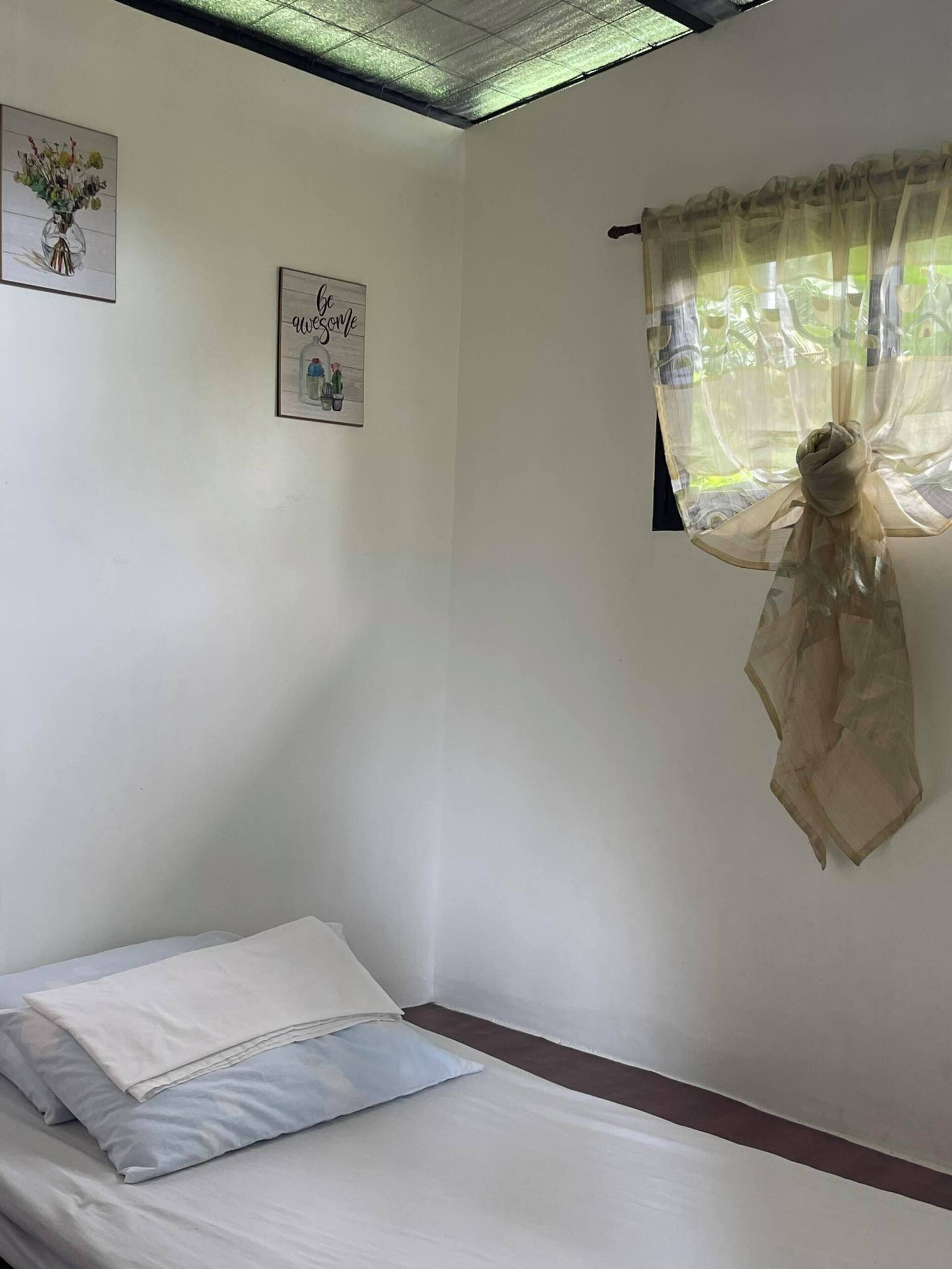 A single bed with white and light blue bedding, a small framed welcome sign, and a window with sheer curtains.