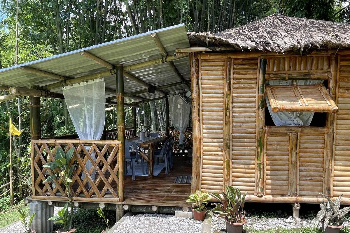 Una cabaña de madera con techo de paja, un porche cubierto y un comedor con mesas al aire libre.