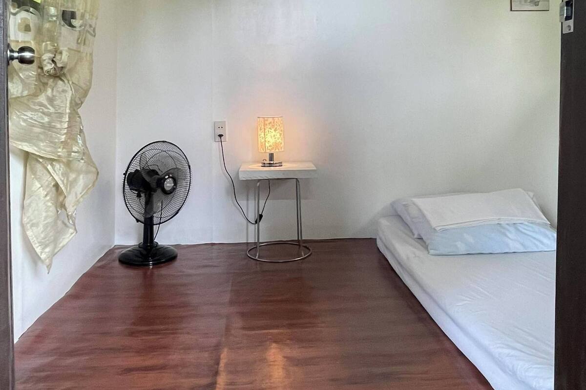 Una habitación con una cama, un ventilador de pie, una mesa con una lámpara y una pared con dos cuadros enmarcados.