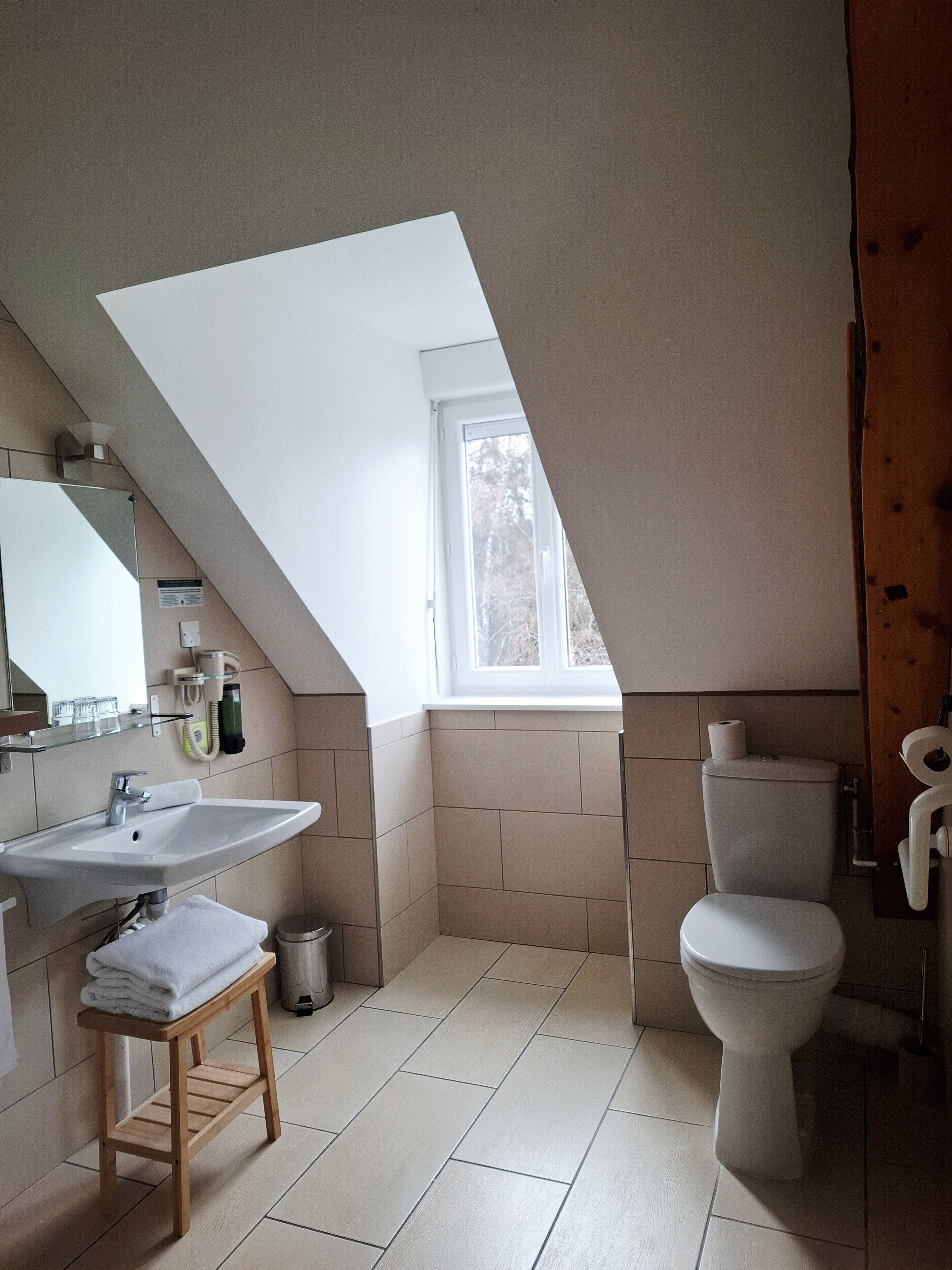 A bathroom with a sloped ceiling, a window, a sink, a toilet, and a small wooden stool.