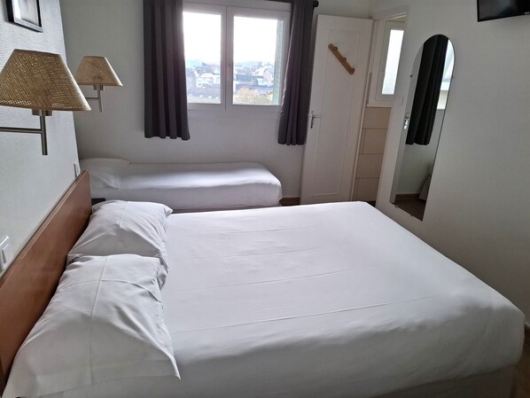 A hotel room with a large bed, a bedside table, a lamp, a window with curtains, and a mirror.