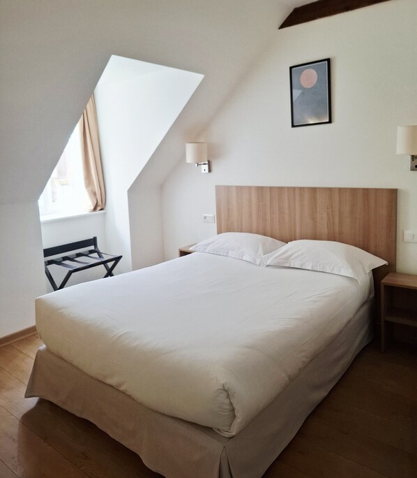 A neatly made bed in a room with a sloped ceiling, a small table, and a wall-mounted lamp.