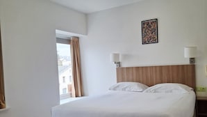 A hotel room with a large bed, two bedside lamps, a headboard, and a window with curtains. - Le grand hôtel ussel (Ussel)