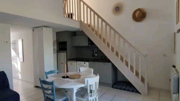 A dining area with a table and chairs, a kitchen in the background, and a staircase leading to an upper level. - House with view of the pines (Narbonne)