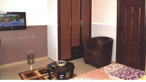 A hotel room with a TV on the wall, a wooden wardrobe, a leather armchair, and a small table with a bowl. - NAPTIN EXECUTIVE HOTEL AND SUITES  (Jos)