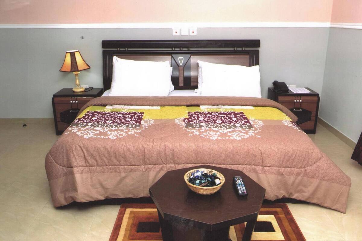 A neatly made bed with a brown bedspread and decorative pillows, a bedside table with a lamp, and a small table with a basket and remote control in front of the bed.