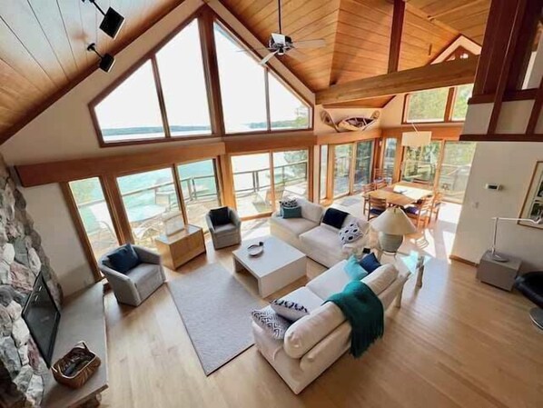 A spacious living area with a high ceiling, large windows, and a stone fireplace.