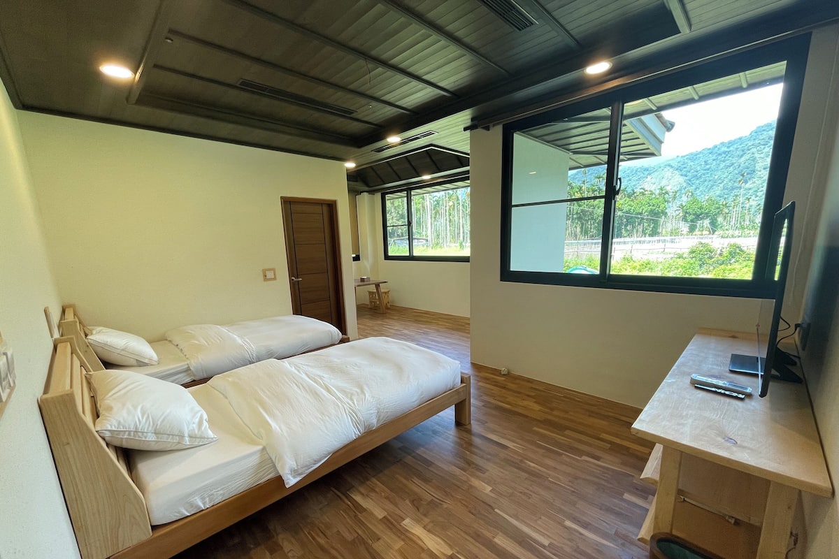 Ein Zimmer mit zwei Holzbetten, einem Schreibtisch mit Computer und großen Fenstern mit Blick auf Grünflächen und Hügel.