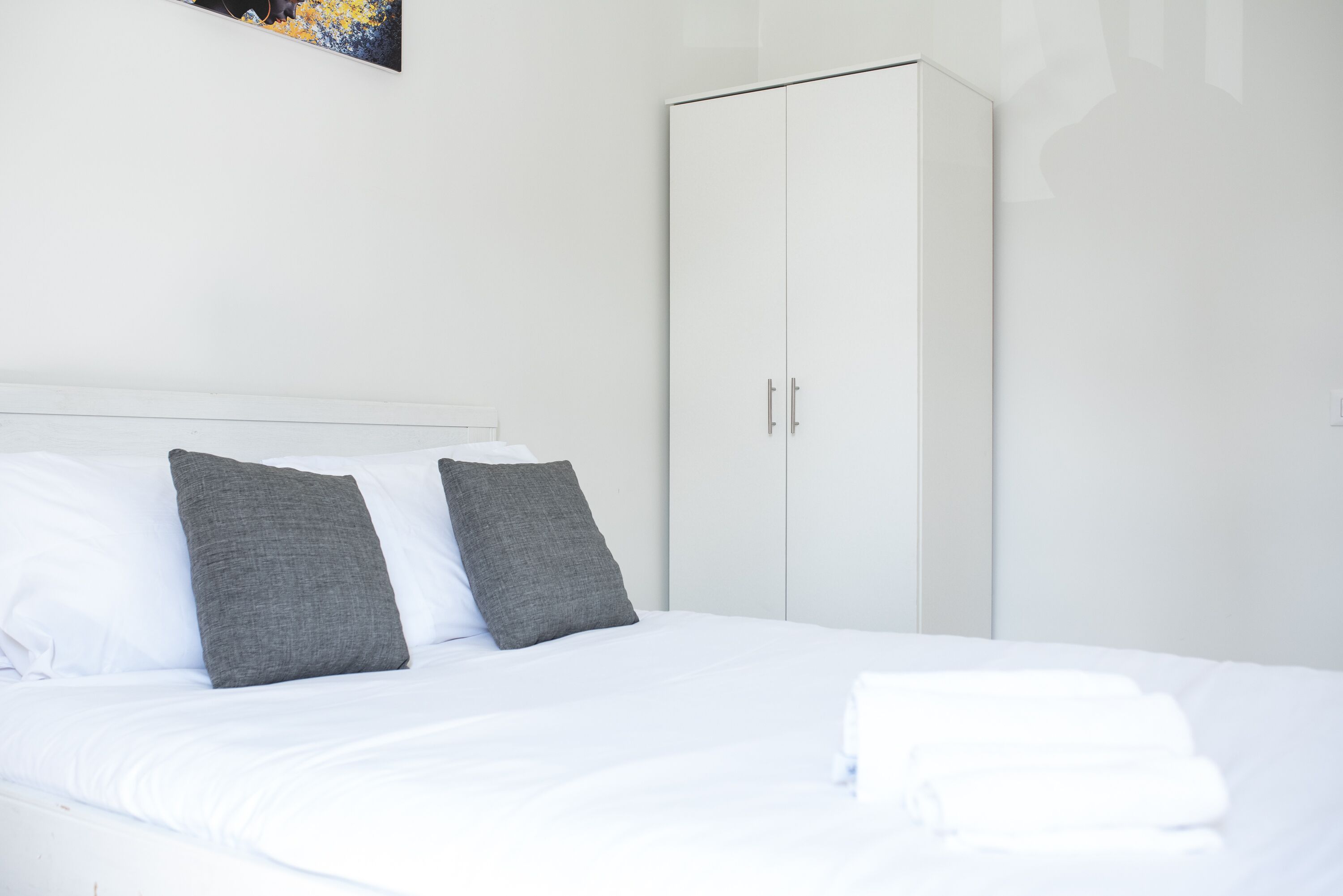Un lit bien fait, avec une literie blanche et deux oreillers gris, une armoire blanche et une œuvre d’art accrochée au mur.