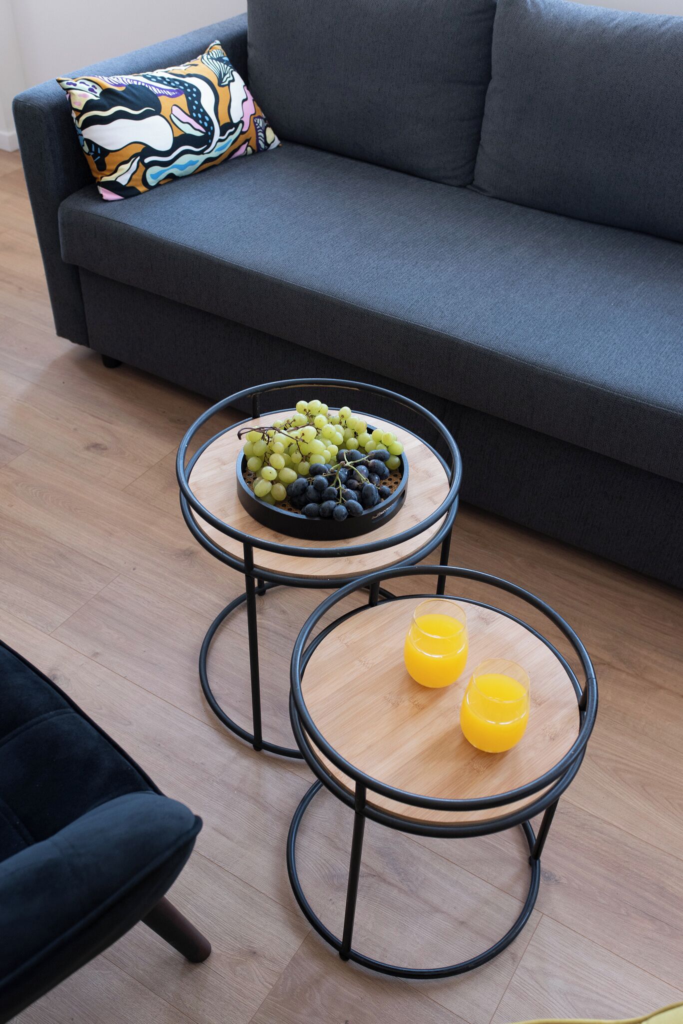 A modern living room with a grey sofa, a colorful pillow, two round wooden tables with metal frames, one holding a bowl of fruit and the other with two glasses of orange juice.