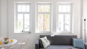 A bright room with a gray sofa, a round dining table with chairs, and three windows with white frames. - PORT CITY HAIFA- Authentic German Colony (Haifa)