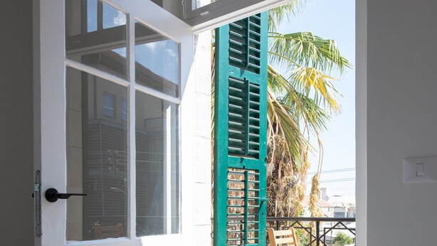 A hotel room with a bed, a window, and a view of a balcony with chairs and palm trees. - PORT CITY HAIFA- Authentic German Colony (Haifa)