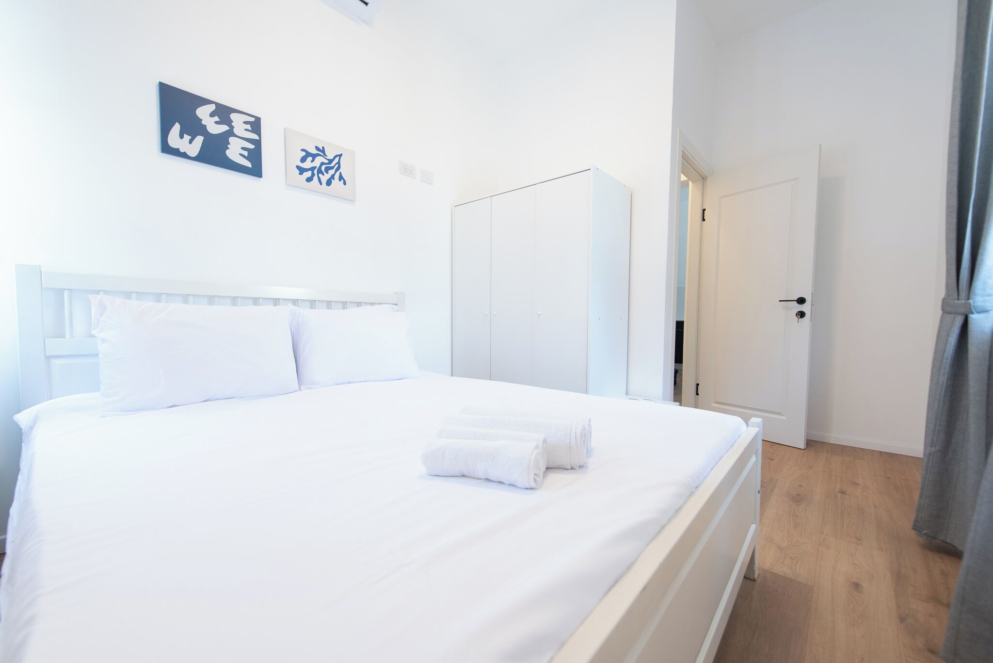 A neatly made bed with white linens and towels, a white wardrobe, and a door leading to another room.