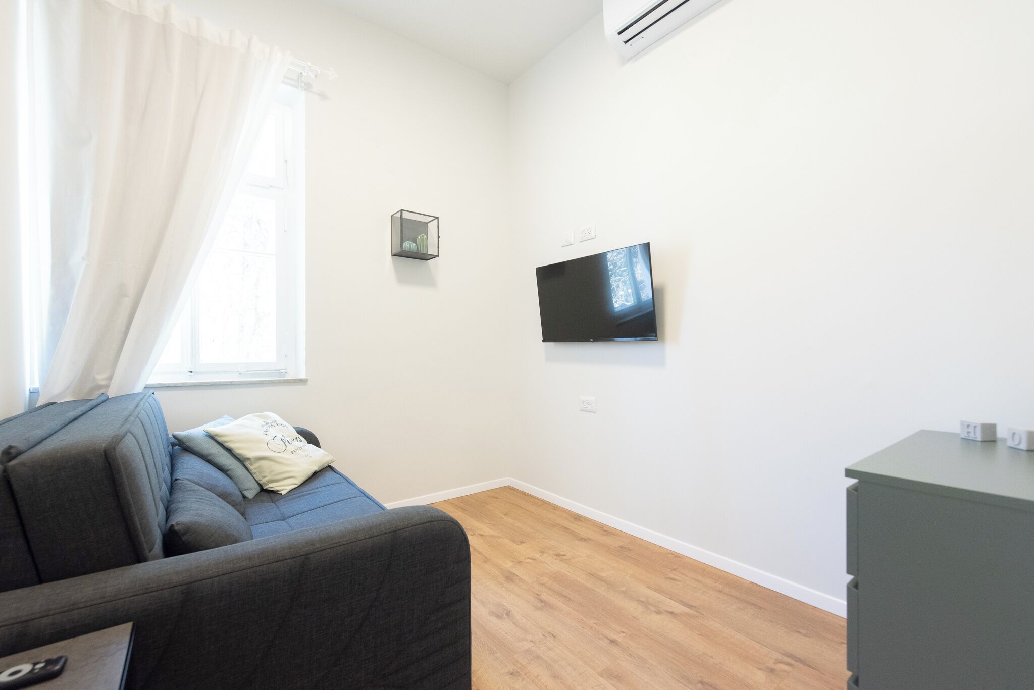 Ein Zimmer mit grauem Sofa, einem flachbildfernseher an der Wand, einer kleinen Ablagefläche mit Dekoration und Holzboden.