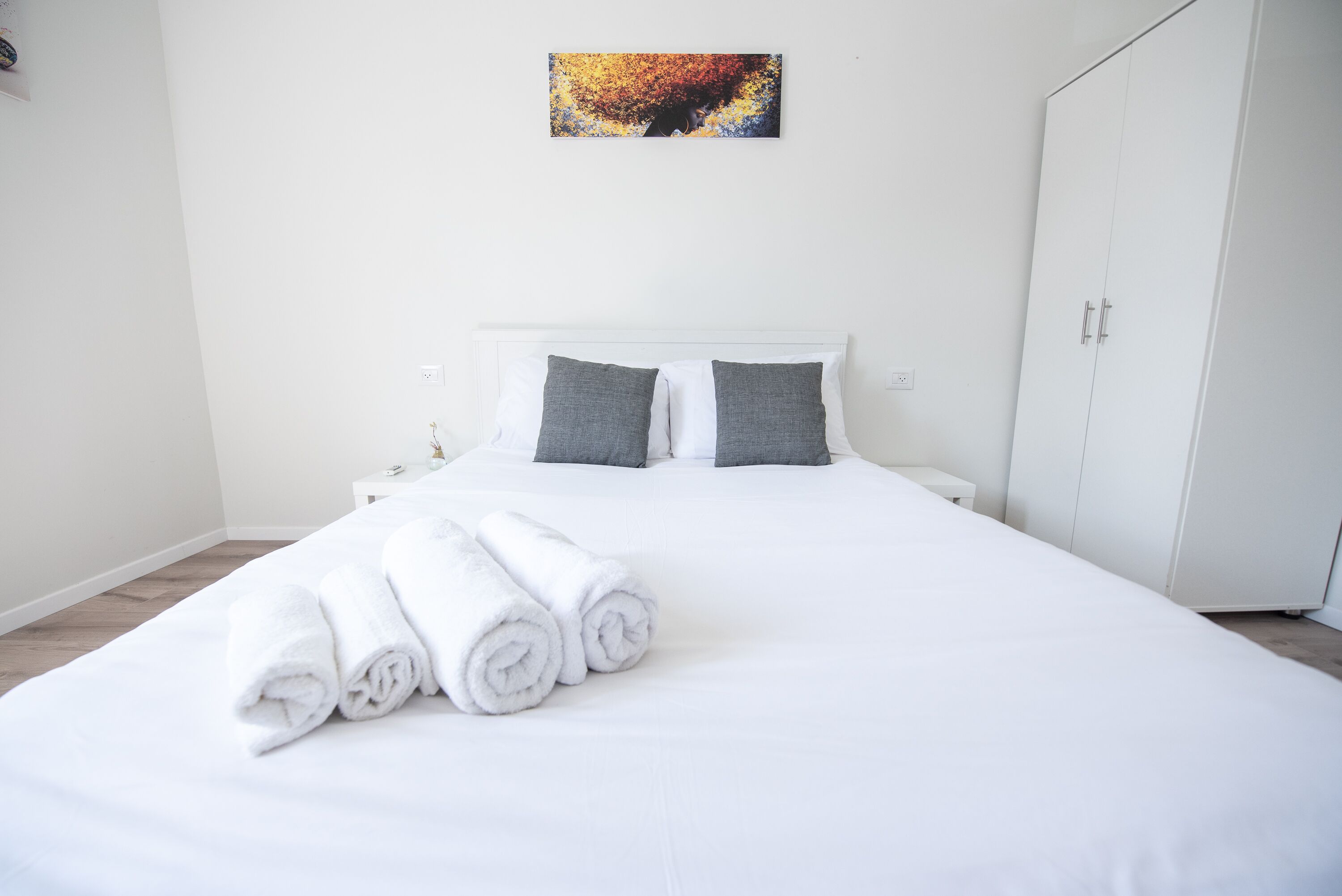 A neatly made bed with white linens and rolled towels, a white headboard, a small bedside table, and a wardrobe.