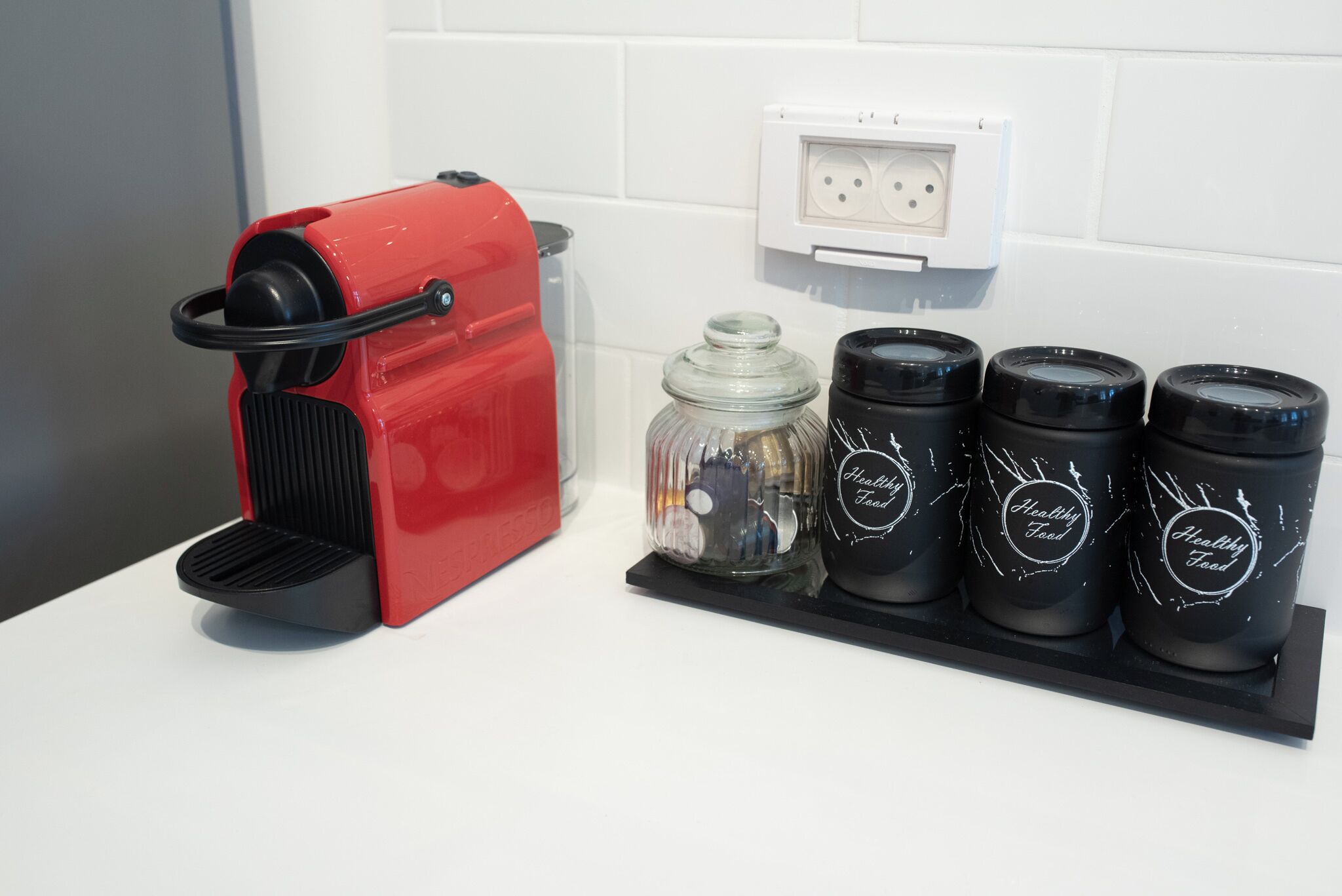 A red coffee maker, a glass jar, and four black mugs with "Healthy Food" labels on a white countertop.