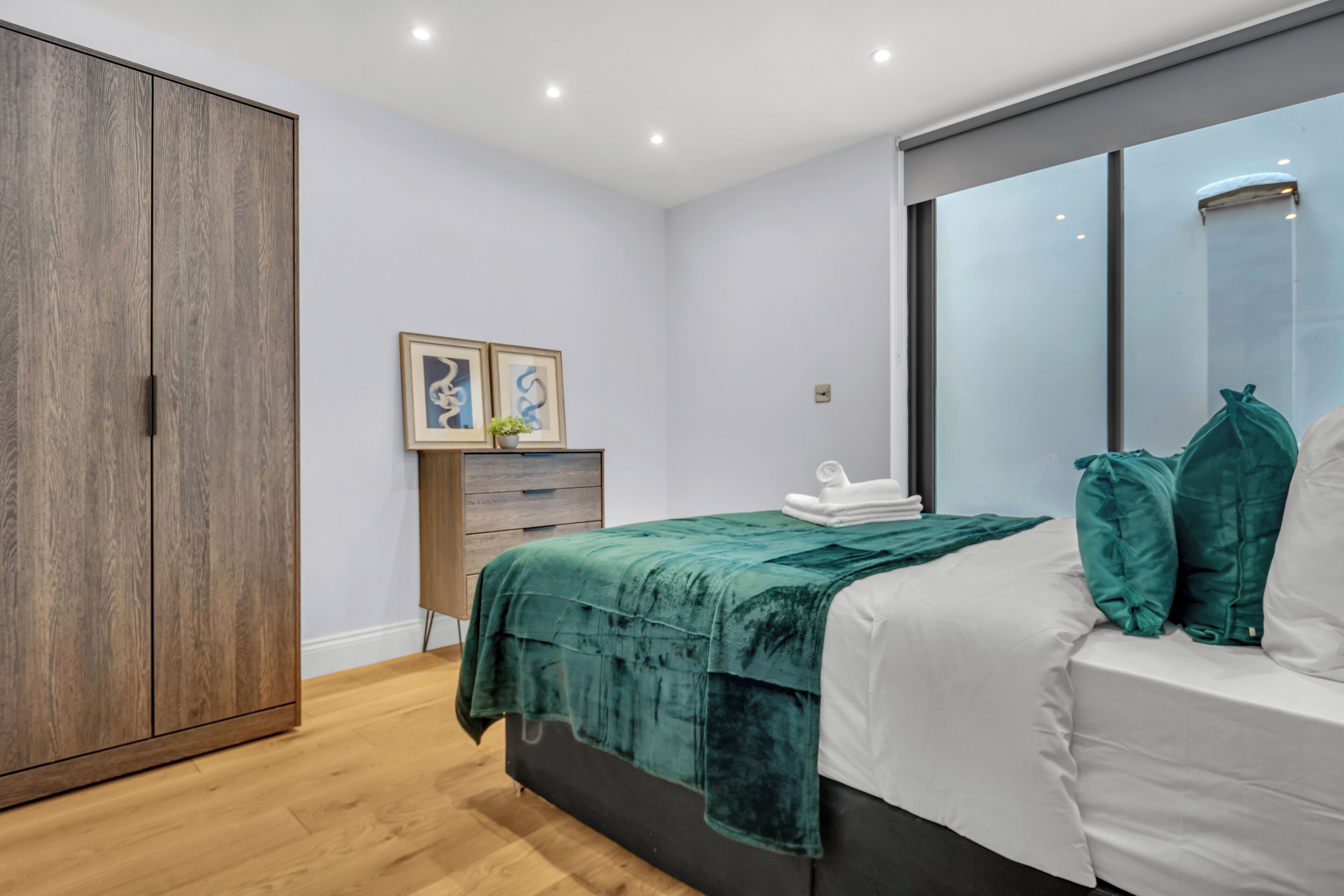 A bedroom with a large wardrobe, a bed with a green blanket, white pillows, and a view of the outside through a window.