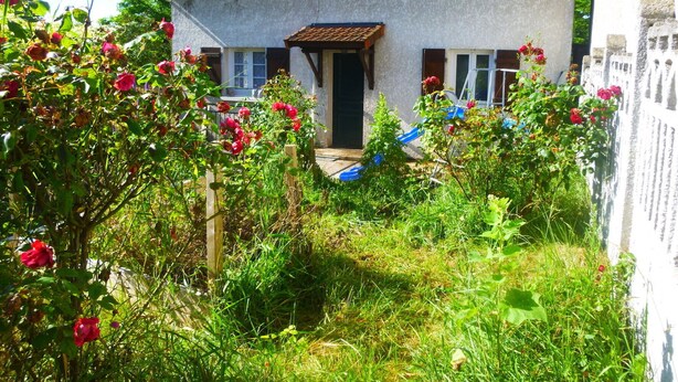 Uma casa com telhado de telhas, porta da frente e um pequeno jardim com flores vermelhas.