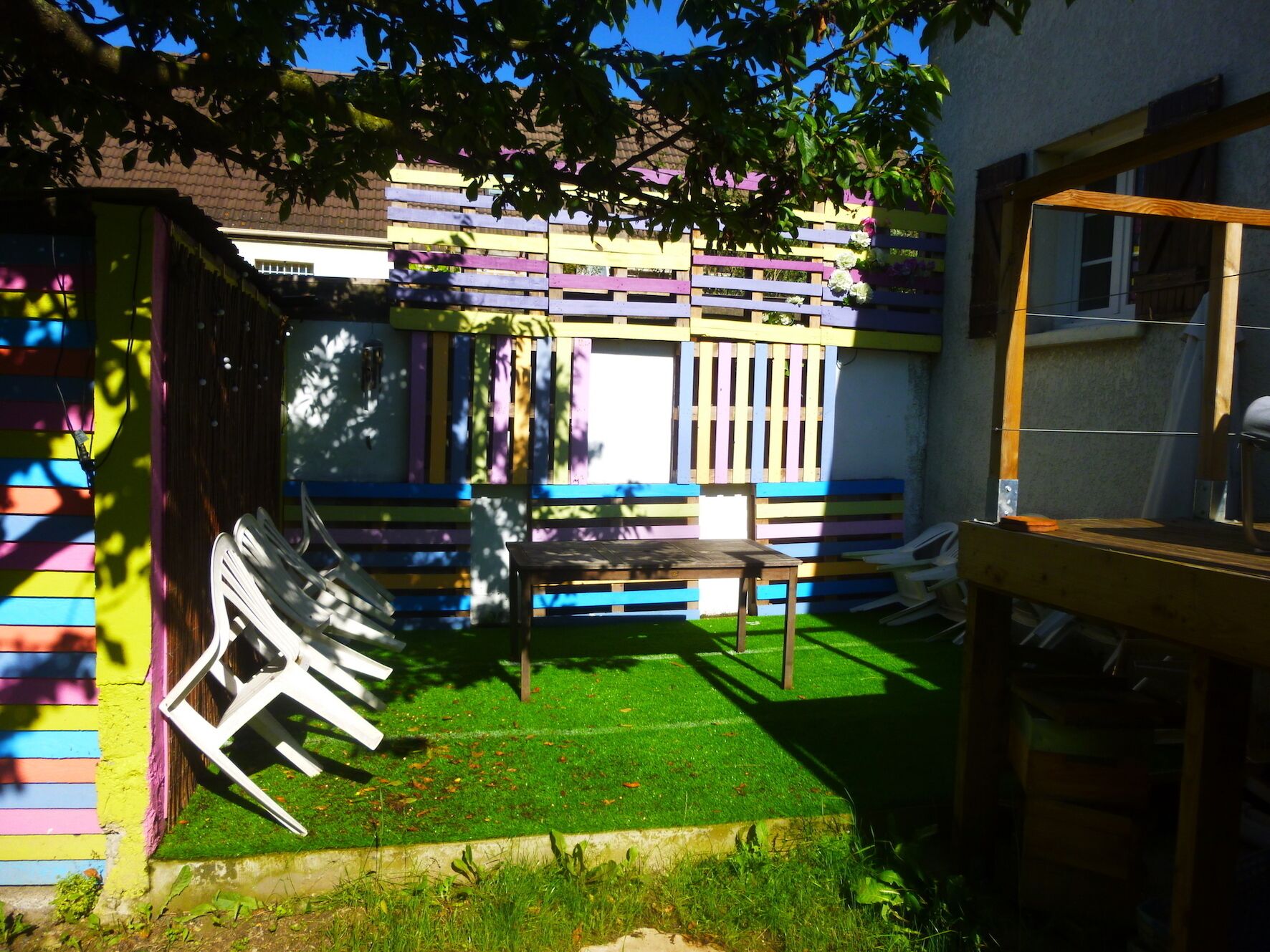 Un área de estar al aire libre llena de color, con una mesa y sillas de madera, rodeada por un cerramiento con paneles multicolores.