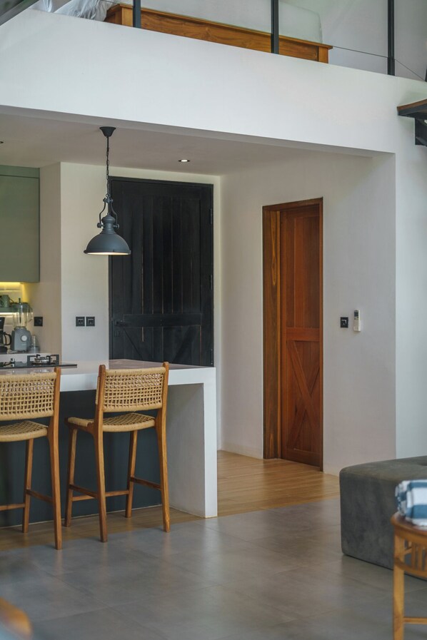 A modern kitchen with a dining area, featuring a wooden door, a black pendant light, and a kitchen counter with a blender. - Loft Valentin (Canggu)