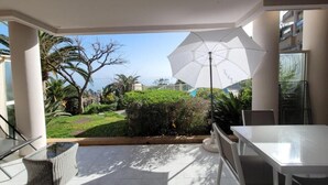 Uma área de varanda coberta com uma mesa e cadeiras brancas, um guarda-sol grande e vista para um jardim bem cuidado e o oceano.