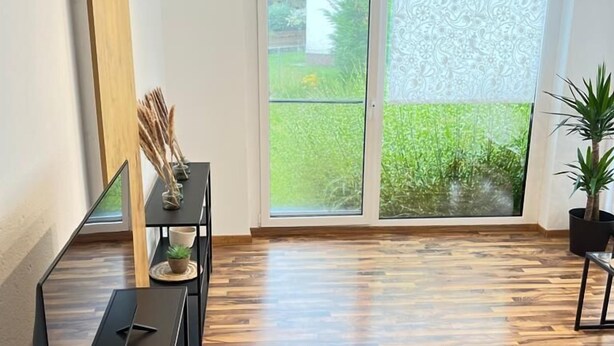 Une pièce spacieuse dotée d’un parquet en bois, d’une porte-fenêtre coulissante donnant sur un jardin et d’un meuble TV noir.