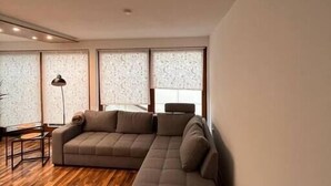 Un salon moderne avec un canapé d’angle, un plancher en bois et de grandes fenêtres ornées de rideaux à motifs.