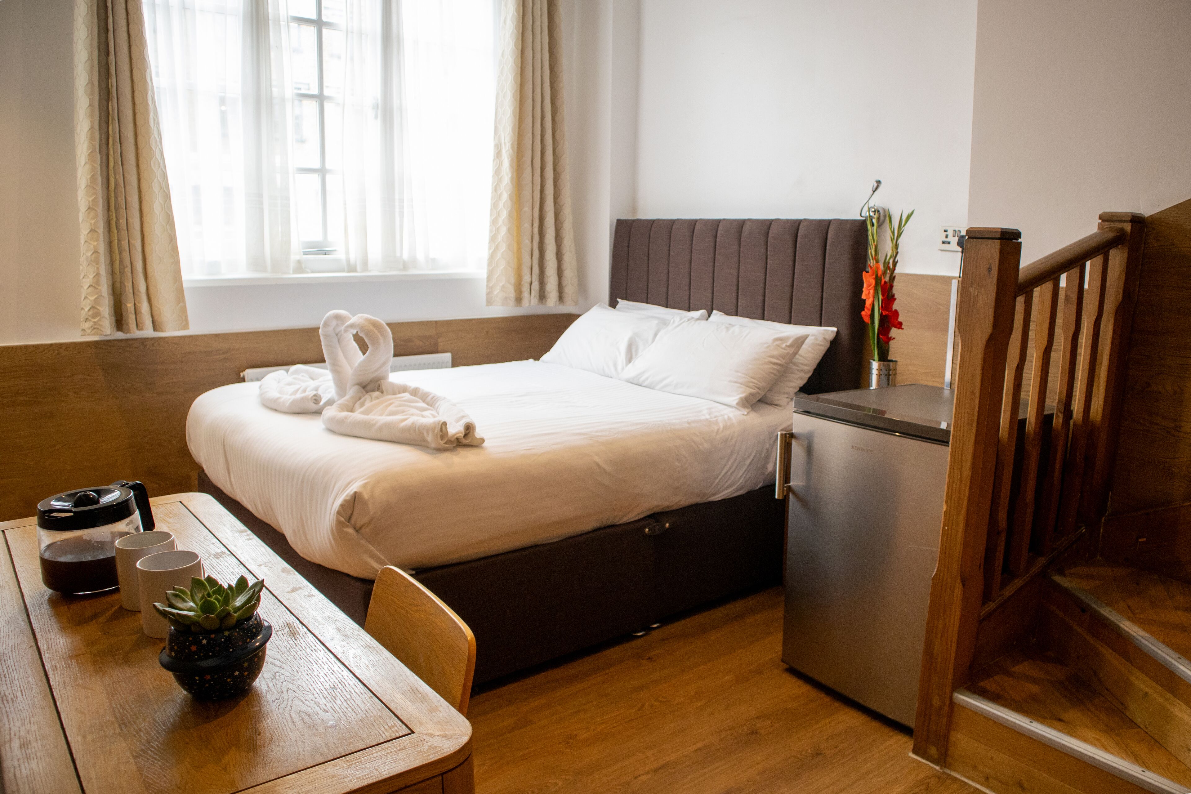 Una habitación de hotel con una cama, una mesita con una planta, un refrigerador y una escalera de madera.